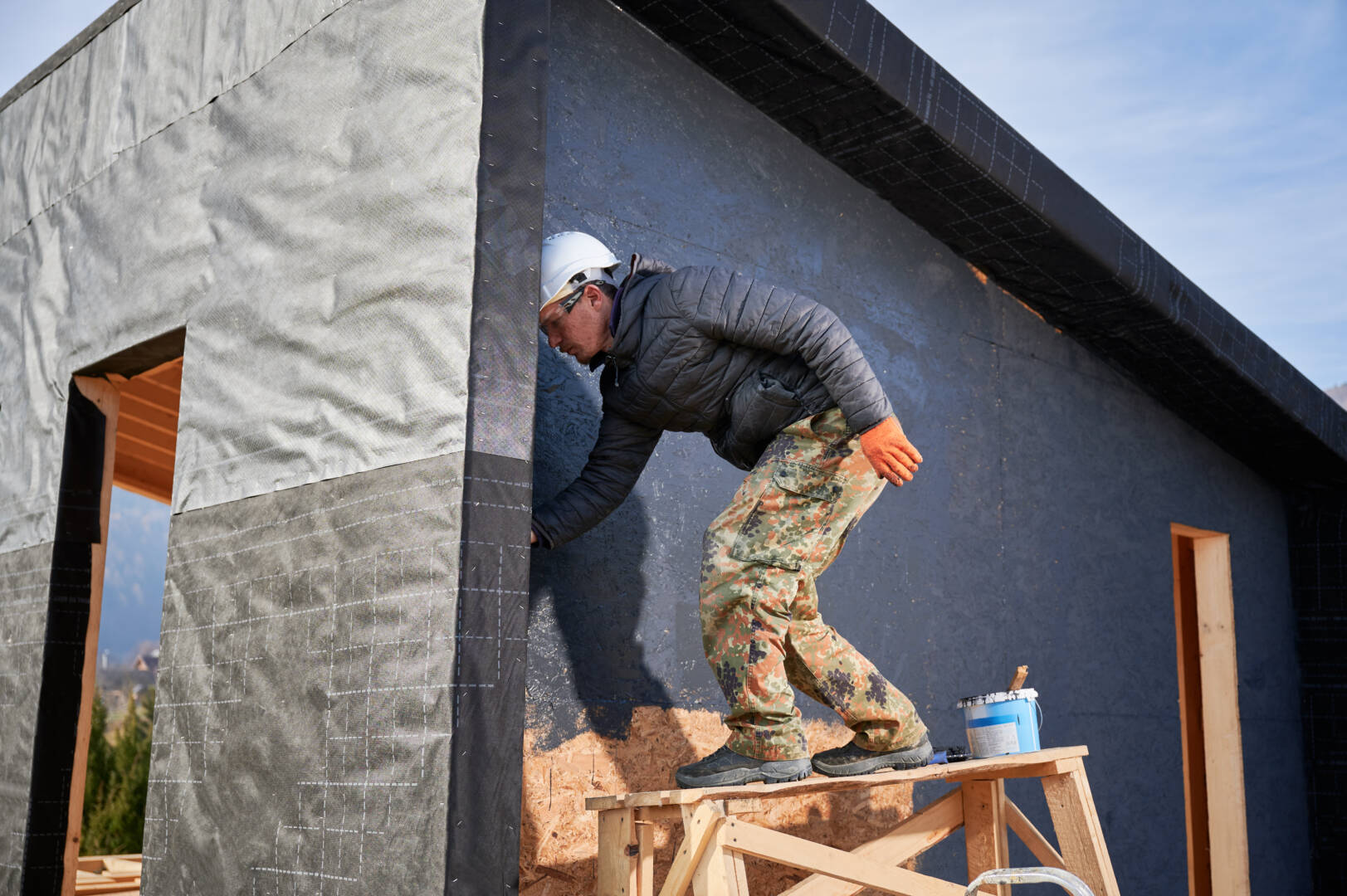 Male painter using paint roller, doing exterior paint work in a black color. Man worker building wooden frame house. Carpentry and construction concept.